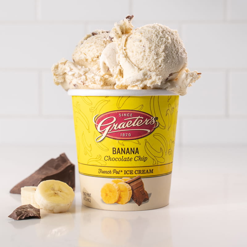 Banana Chocolate Chip: yellow ice cream with light brown chocolate chunks throughout overflowing from a pint with banana slices and chocolate chunks next to it in front of a white background