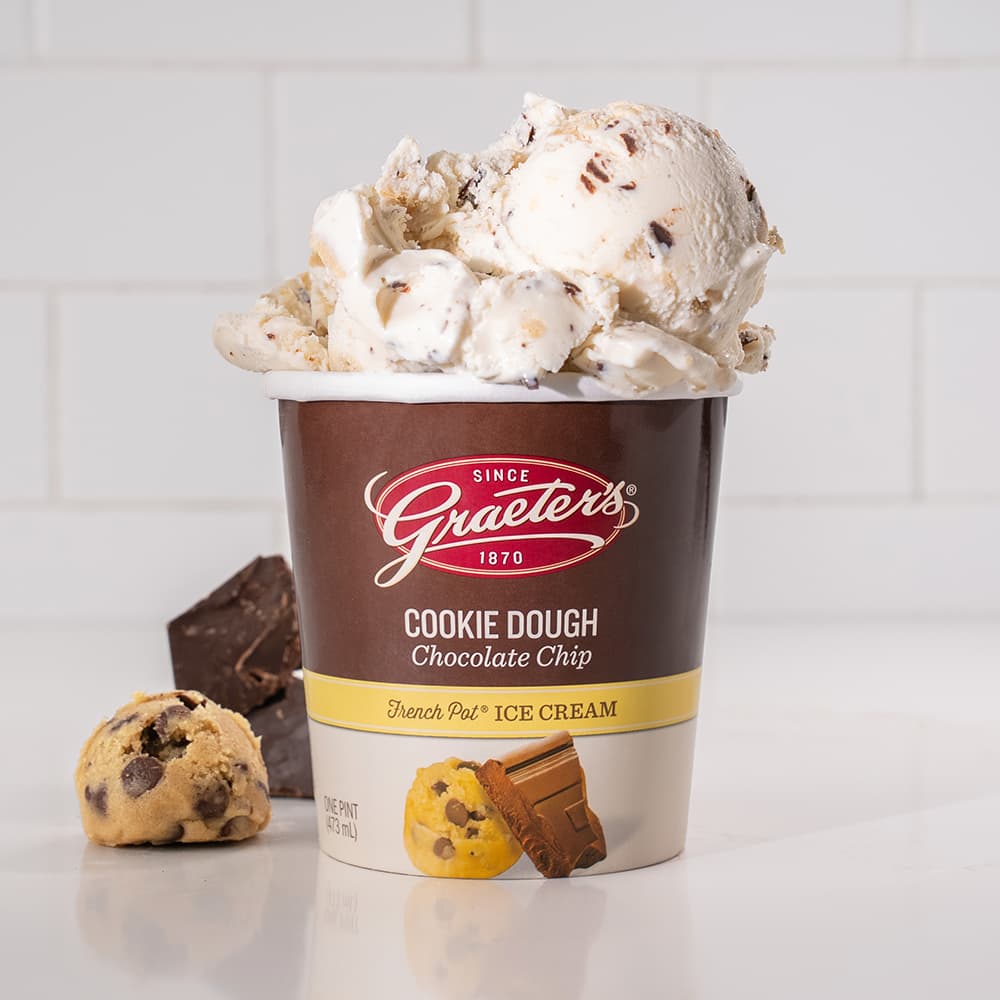 Cookie Dough Chocolate Chip ice cream: white ice cream with dark chocolate chips and cookie dough balls overflowing from the pint. Next to it is a chocolate chip cookie dough ball and chocolate chip chunks in front of a white background
