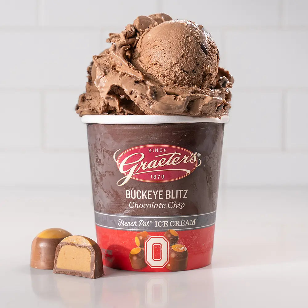 Graeter's Buckeye Blitz Chocolate Chip Ice Cream: dark brown ice cream with balls of light brown peanut butter cookie dough and dark chocolate chips mottled throughout overflowing from a pint with buckeye candy next to it in front of a white background