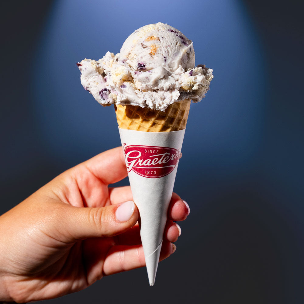 Single dip of Elena's blueberry pie ice cream: a light purple ice cream with blueberries and pie crust within on a sugar cone