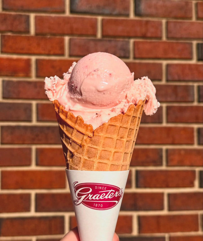 a single scoop of pink strawberry ice cream in a waffle cone in front of a brick background