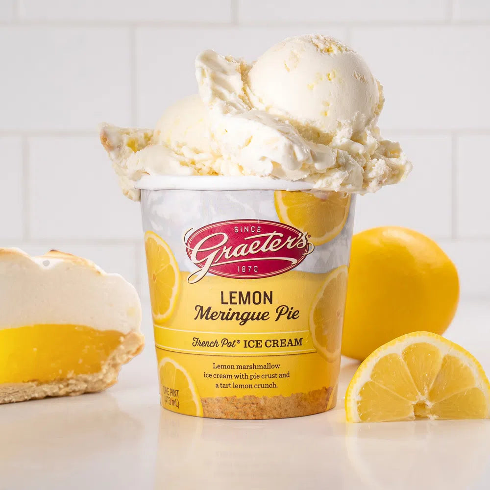 a Graeter's Lemon Meringue Pie ice cream pint overflowing with yellow white ice cream with light brown graham cracker pieces throughout. Next to the pint are lemons and a bright lemon meringue pie in front of a white background