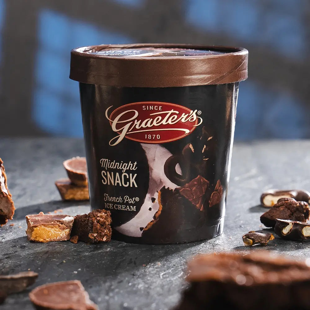 Graeter's Midnight Snack ice cream pint. It is a dark pint with a crescent moon on it, it is in front of a dark background with peanut butter cups, brownies, and chocolate covered pretzels scattered around it