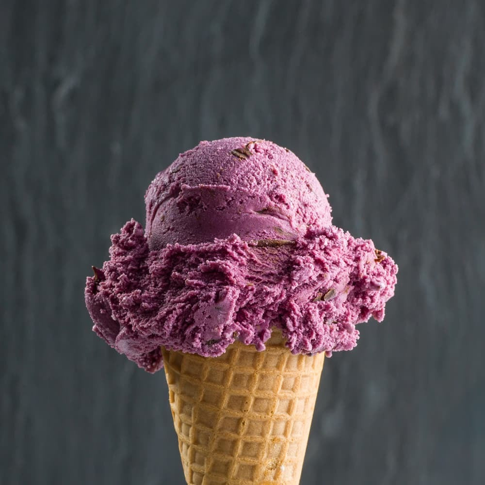 a perfect single scoop cone with dark purple black raspberry chocolate chip ice cream in front of a dark grey background
