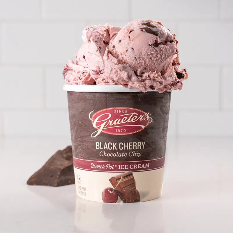 Graeter's Black Cherry Chip: pink ice cream with chocolate chunks and cherry pieces throughout overflowing from a pint, chocolate chunks are sitting next to it in front of a white background
