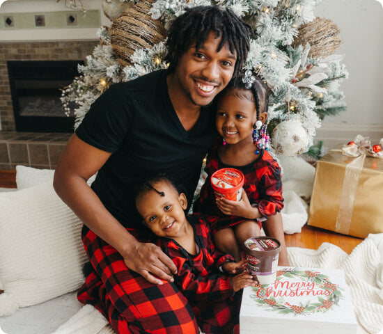 Family of three in matching red plaid pajamas sitting on a couch with a Christmas tree and presents in the background.