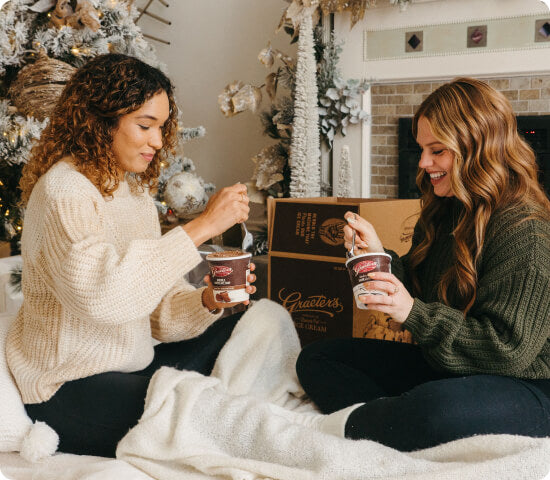 Two women sitting on a bed with a Christmas tree and fireplace in the background, holding Graeter's Ice Cream.