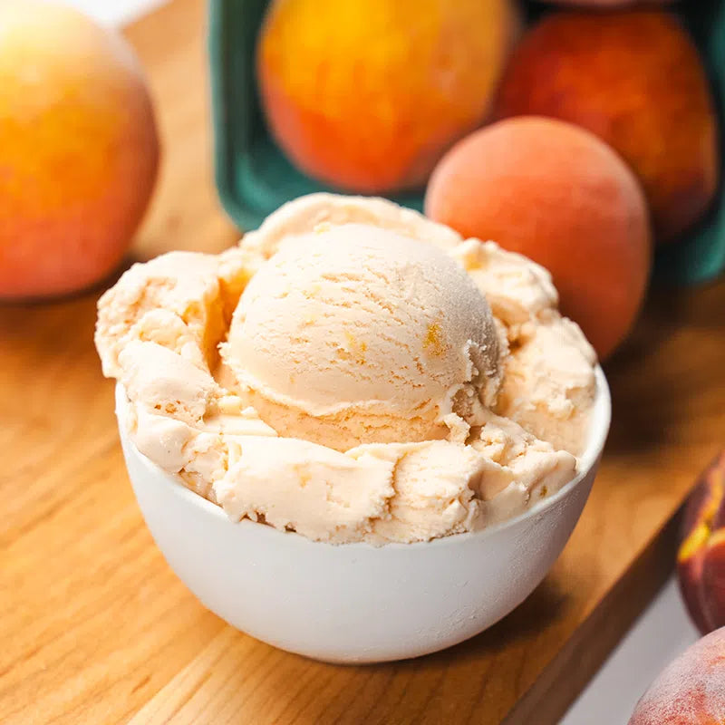 Peach ice cream: peachy orange ice cream with peach chunks peeking through in a white bowl with peaches in the background on a wooden cutting board