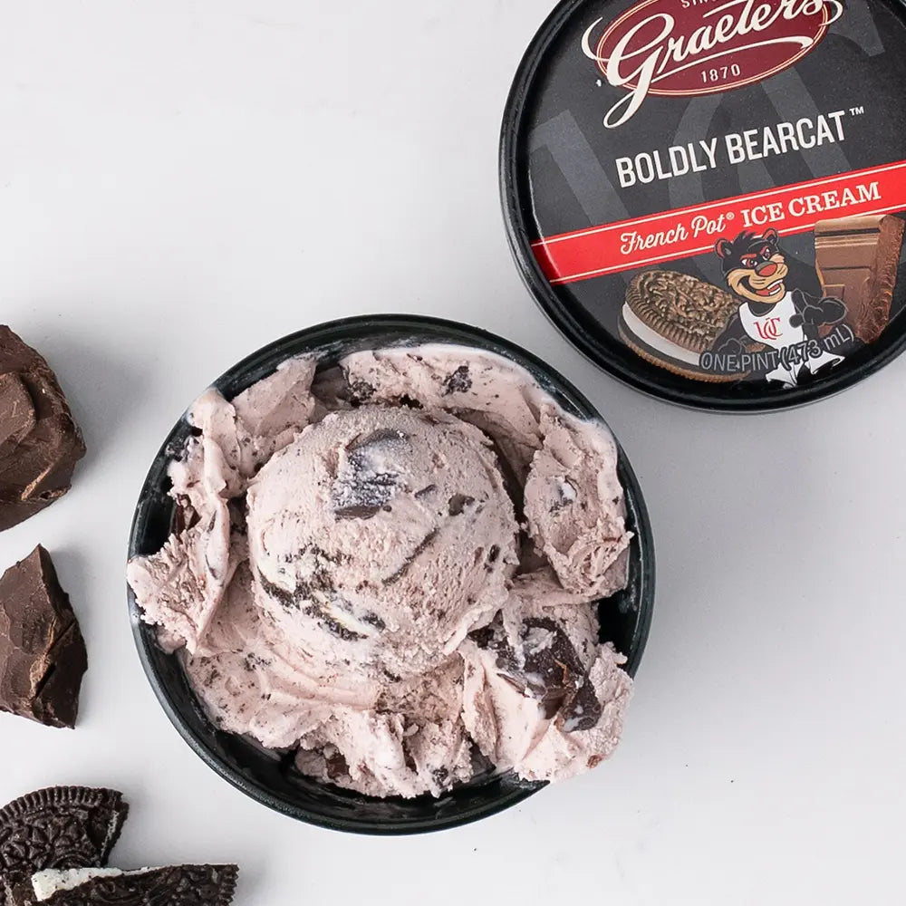 Graeter's Boldly Bearcat: white pink ice cream with chocolate chunks and cookie pieces scattered within it in a black bowl. Next to it are cookie pieces, chocolate chunks, and a pint lid on a white background