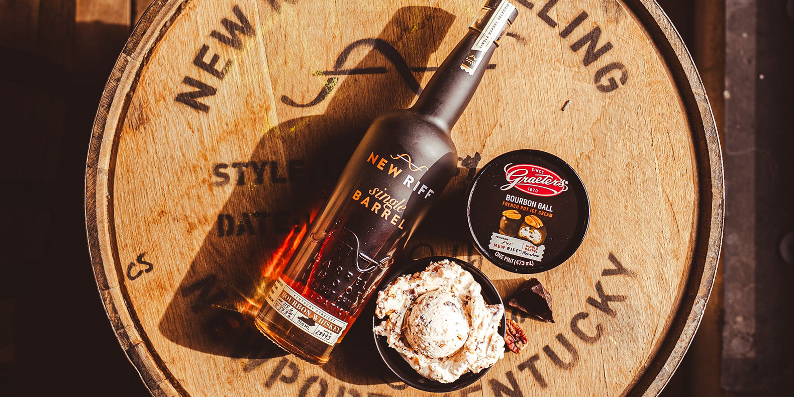 brown ice cream with chunks of pecan sitting next to a bottle of New Riff bourbon and the Graeter's Bourbon Ball ice cream on top of a barrel of bourbon